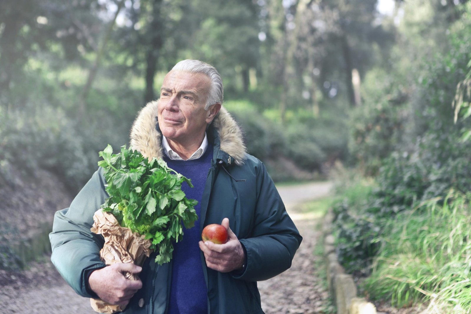 Elderly man enjoying nature, holding fresh produce on a scenic pathway.